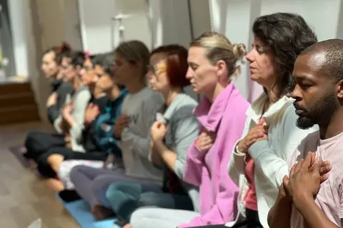 A diverse group of people sitting in a row with hands on chests during a meditation session.