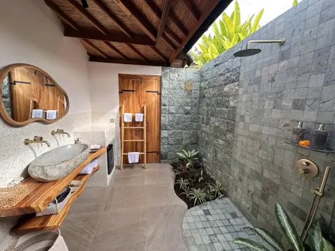 An outdoor tropical bathroom with a stone vessel sink, wooden vanity, and grey stone-tiled shower.