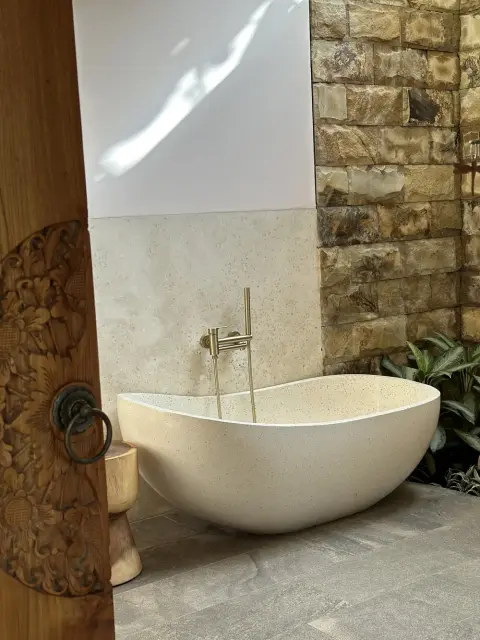 A cream-colored stone bathtub in an outdoor bathroom with a textured stone wall.