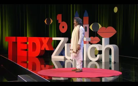 TEDxZurich speaker in white traditional attire on a stage with large red and white lettering.